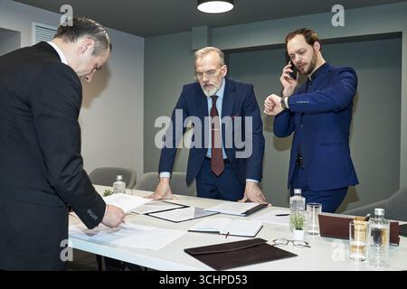Three Caucasian men, including middle aged and senior, standing around conference table reviewing documents, one middle aged man talking on smartphone and checking wristwatch Stock Photo