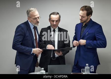 Three middle aged Caucasian men standing together discussing documents in office setting, one man holding folder while others listening attentively, laptop and water bottles on table Stock Photo
