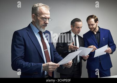 Three middle aged Caucasian men standing together reviewing documents, two colleagues discussing paperwork while third executive reading sheet, business professionals collaborating in office setting Stock Photo