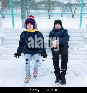 Two children, a boy and a girl, dressed warmly in winter coats and hats, sit side-by-side on a white bench covered in snow. The serene winter scene pr Stock Photo