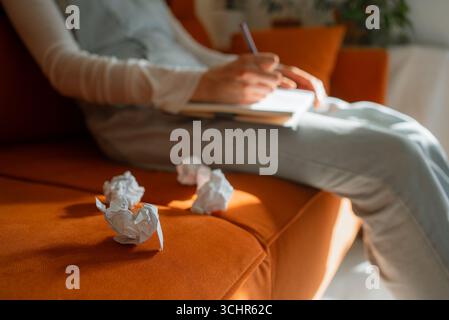 Woman sitting on an orange sofa is writing on a notebook with a pen, several crumpled pieces of paper lying next to her, suggesting creative writing or brainstorming Stock Photo