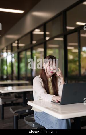 Focused Asian woman works on laptop at modern cafe. Remote worker studies business project in a bright collaborative space. Stock Photo