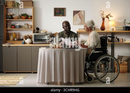 Senior Caucasian woman with disability sitting in wheelchair discussing documents with young adult Black man at table in home kitchen, both focused on paperwork and conversation Stock Photo