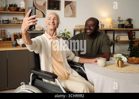 Senior Caucasian woman in wheelchair holding smartphone taking selfie with young Black man sitting beside her at table smiling, both looking at phone screen indoors Stock Photo