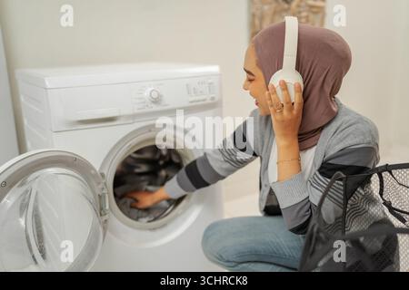 A woman in headphones operates a washing machine, enjoying music while doing chores Stock Photo