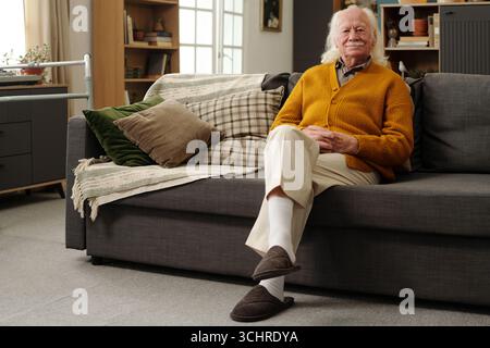 Portrait of senior Caucasian man sitting on sofa crossing legs, smiling gently while looking slightly away from camera, showing relaxed posture in modern living room setting Stock Photo