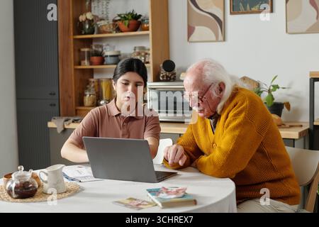 Young adult Hispanic woman assisting senior Caucasian man using laptop at kitchen table, both focusing on screen while discussing documents and working together on paperwork Stock Photo