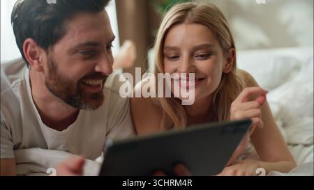Family laughing Caucasian couple pair typing tablet shopping online in bedroom morning married woman discussing app wife man using technology Internet Stock Photo