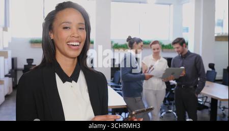 Smiling woman wearing blazer checking smartphone at office with team reviewing documents and tablet Stock Photo