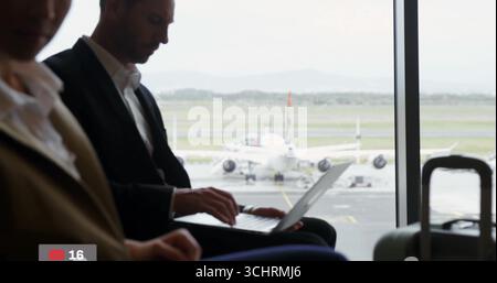 Typing businessman sitting in airport departure lounge, with laptop, suitcase beside large window Stock Photo