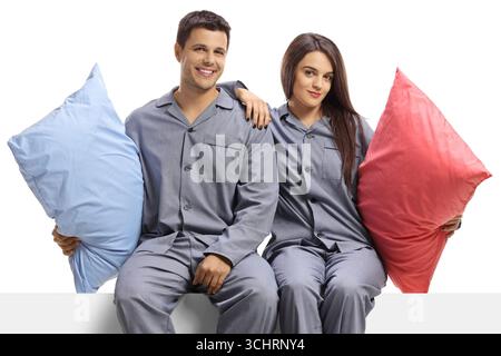 Couple in matching pajamas sitting on a white panel and holding pillows isolated on white background Stock Photo