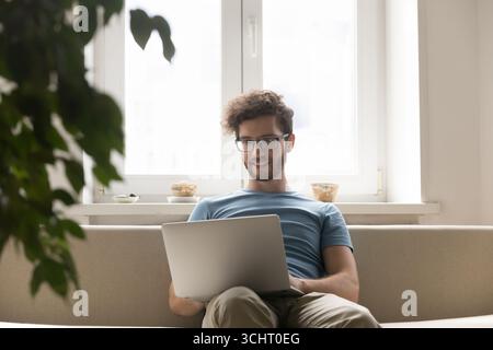 Smiling young guy using notebook on sofa in living room Stock Photo