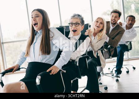 Group of smiling coworkers having fun racing in office chairs during a team bonding activity in a modern workplace Stock Photo