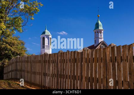 A tall wooden fence  allows only the view of two churches bell towers under blue skies in downtown historical Old Salem, North Carolina, USA Stock Photo