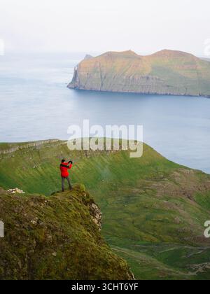Man In Red Jacket Standing On Rocky Cliff Taking Photo Of Scenic Vagar Island In Faroe Islands Stock Photo