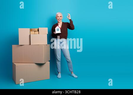 Cheerful mature woman on a blue background holding a model house next to stacked cardboard boxes in casual attire Stock Photo