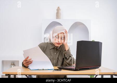 A Focused Young Woman Dressed in Hijab Engaged in Serious Study with Papers and Laptop Stock Photo