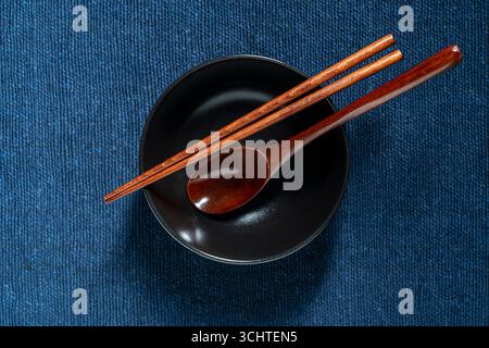 A pair of wooden chopsticks and spoon lying across an empty black soup bowl on blue fabric background, close up, top view Stock Photo