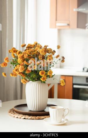 Autumn cozy home interior with coffee in white cup. Beautiful bouquet of orange flowers in vase at modern kitchen. Season cozy decoration at home. Clo Stock Photo
