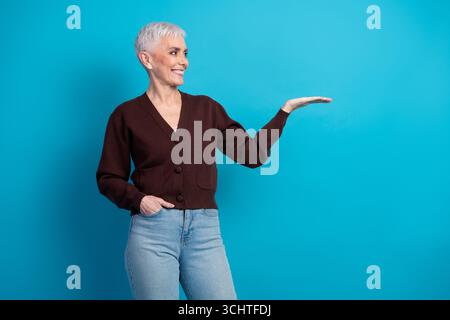Confident mature woman in casual outfit smiling and gesturing with hand on blue background demonstrating space Stock Photo