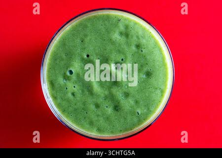 Glass of green smoothie on a red background, close up, top view. Healthy organic drink made from banana, dill, parsley and avocado. Bubbles and foam o Stock Photo