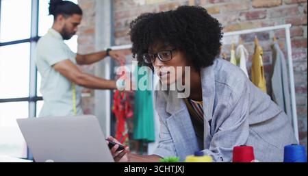Female fashion designer leaning on desk holding stylus in loft studio, with laptop and tape measure Stock Photo