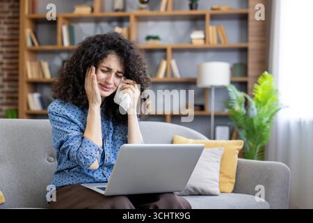 Sad woman is crying while working with her laptop on the sofa at home, wiping her tears with a tissue, having some problems with her online business activity Stock Photo