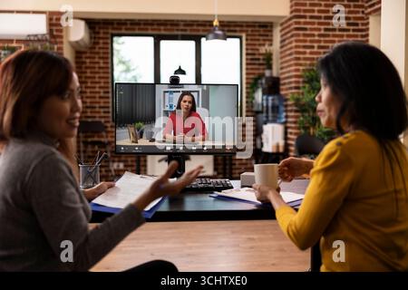 Asian women with clipboards, having videoconference call with caucasian manager on computer, reviewing cv resumes of job candidates. Female startup team utilizing digital communication for a meeting. Stock Photo