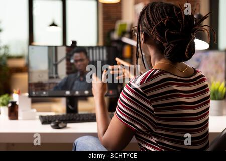 Black woman uses desktop pc for virtual meeting, discussing marketing strategies with business advisor from home. Male consultant appears on screen, offering strategic insights to focused entrepreneur Stock Photo
