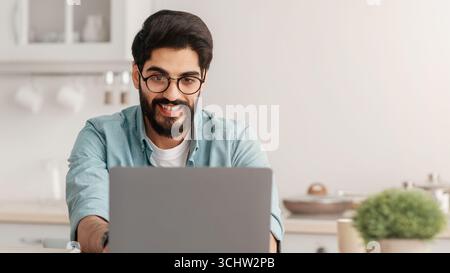 Remote business. Happy arab freelancer man working with laptop in kitchen, typing on computer, enjoying online job Stock Photo