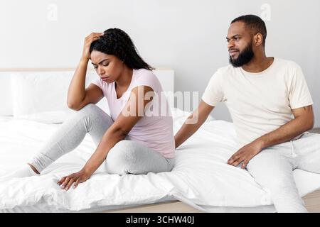 Serious young black man calm to sad desperate depressed wife, sit on bed in bedroom interior Stock Photo