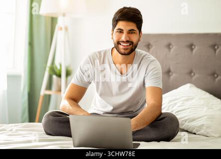 Freelance career. Happy arab man sitting on bed and using laptop computer, working remotely from home Stock Photo