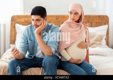 Young pregnant muslim woman offended to her husband busy with smartphone, arab lady in hijab feeling lonely Stock Photo