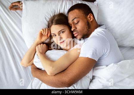 Young black guy sleeping and hugging his beautiful Caucasian girlfriend in bed, top view Stock Photo