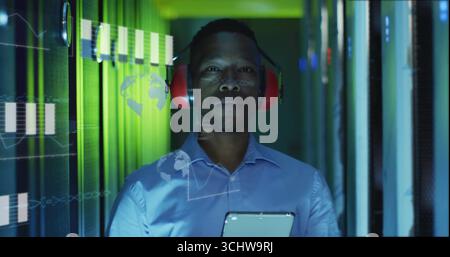 Inspecting racks tech wearing blue shirt earmuffs holding tablet in data center, with overlays Stock Photo