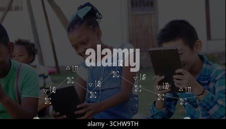 Smiling girl in denim overalls and teal bow tapping tablet on grassy lawn, with friends Stock Photo
