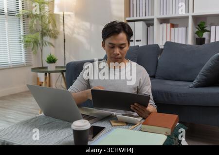 A young man is sitting on the floor at home, checking documents while working with his laptop. He appears focused and engaged, managing his tasks in a Stock Photo