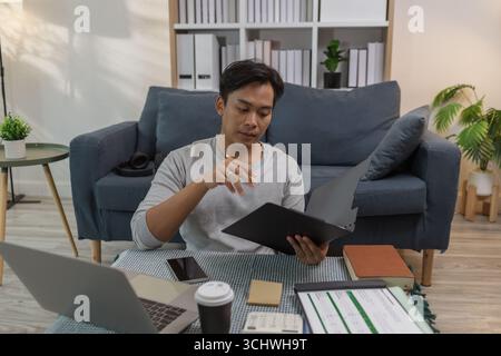 A young man is sitting on the floor at home, checking documents while working with his laptop. He appears focused and engaged, managing his tasks in a Stock Photo
