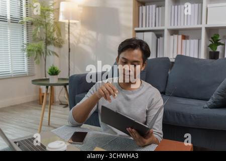 A young man is sitting on the floor at home, checking documents while working with his laptop. He appears focused and engaged, managing his tasks in a Stock Photo