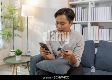 A young man is sitting comfortably on sofa, smiling while using his smartphone and holding a credit card, making an online payment. The scene reflects Stock Photo
