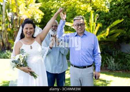 Diverse couple celebrating wedding garden with senior friends, holding hands and smiling Stock Photo