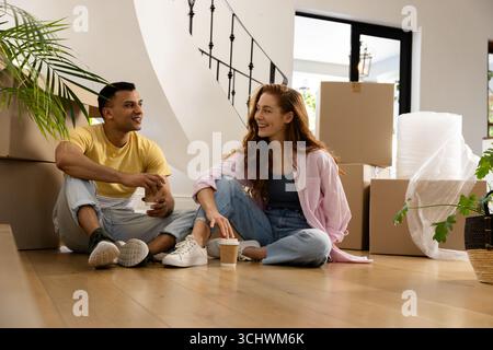 Young diverse couple sitting on floor, surrounded by moving boxes, smiling and talking, at home Stock Photo