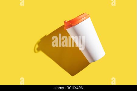 Floating white coffee cup with orange lid on yellow background casting shadow. Stock Photo