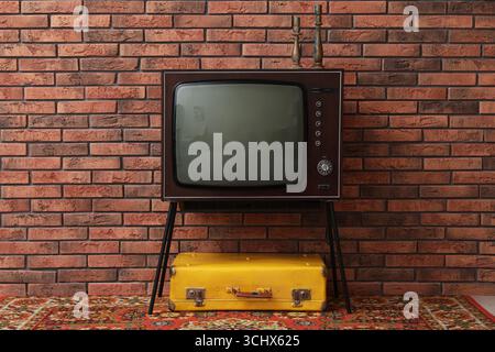 Retro TV on stand, suitcase and carpet near red brick wall. Used stuff Stock Photo