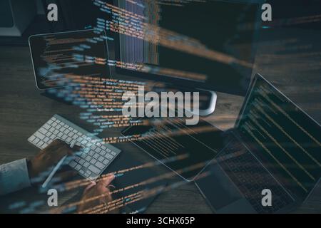 Coding and software development concept, with programmer typing on keyboard surrounded by laptop, tablet, and monitor showing glowing source code line Stock Photo