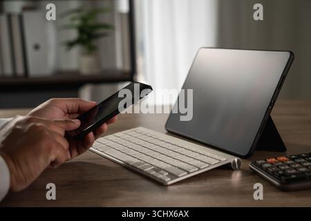 Remote freelance work and digital nomad concept, with person using smartphone at home desk next to tablet, keyboard, and office supplies Stock Photo