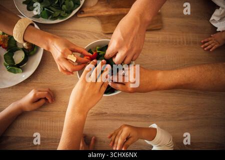 Hands of various ages and ethnicities are reaching into a bowl of fresh salad on a wooden table, emphasizing togetherness and healthy lifestyle choice Stock Photo