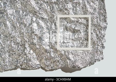Crumpled silver foil background with white frame overlay on a light surface for design template. Stock Photo