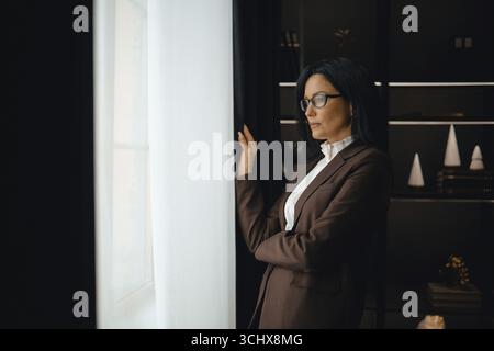 A professional woman dressed in a brown suit stands thoughtfully by a window, gazing outside Stock Photo