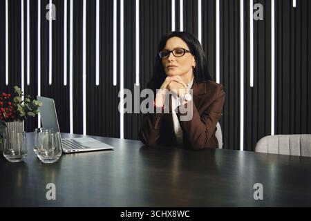 A thoughtful woman with glasses sits at a modern office desk. She has long dark hair and is dressed in a brown suit Stock Photo